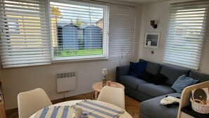 Un salon chaleureux avec un canapé gris, une table ronde recouverte d’une nappe à rayures et une fenêtre avec des stores.