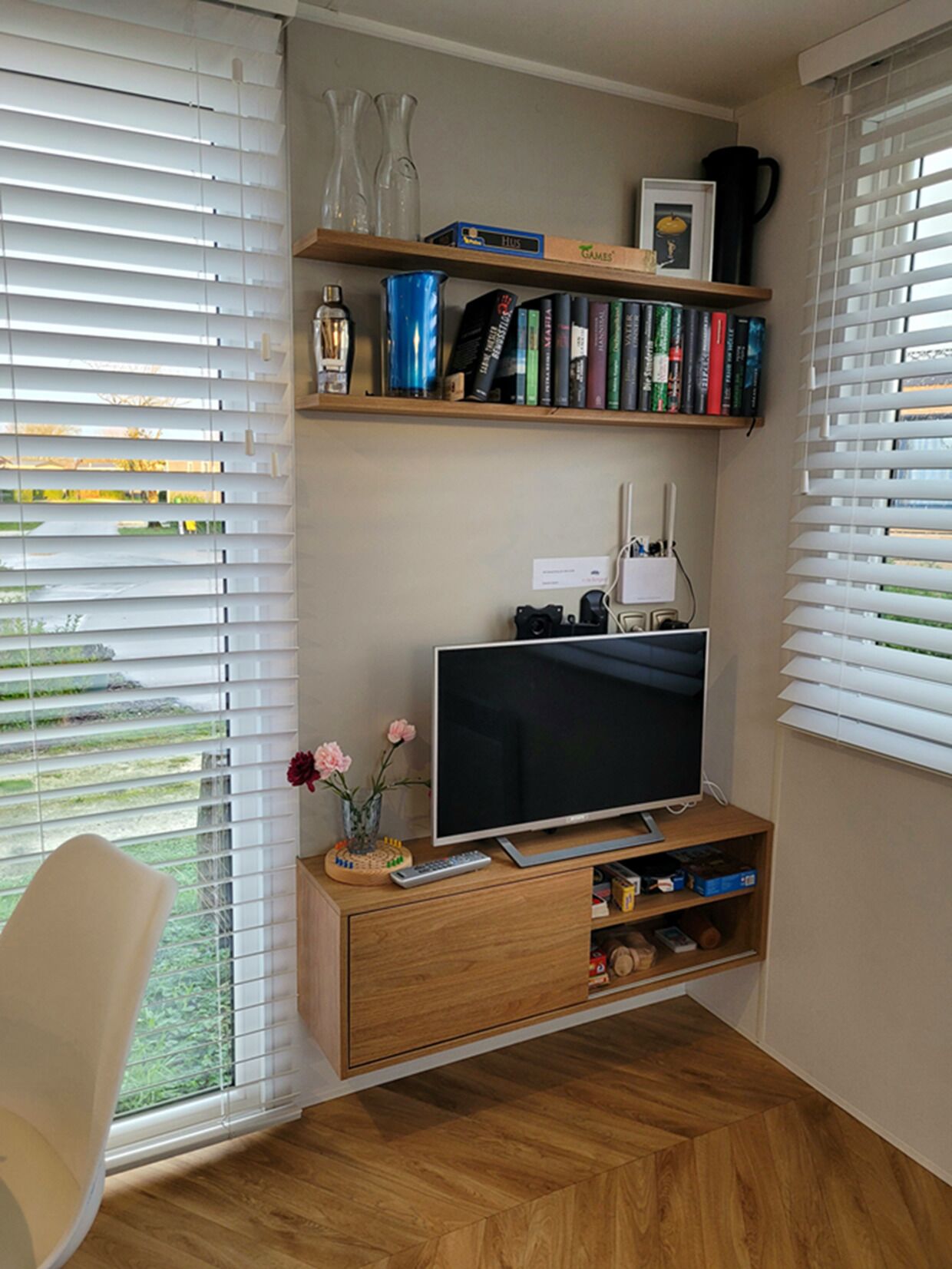 Een moderne woonkamer met een flatscreen televisie op een houten standaard, een boekenkast met boeken en decoratieve objecten, en een raam met jaloezieën.