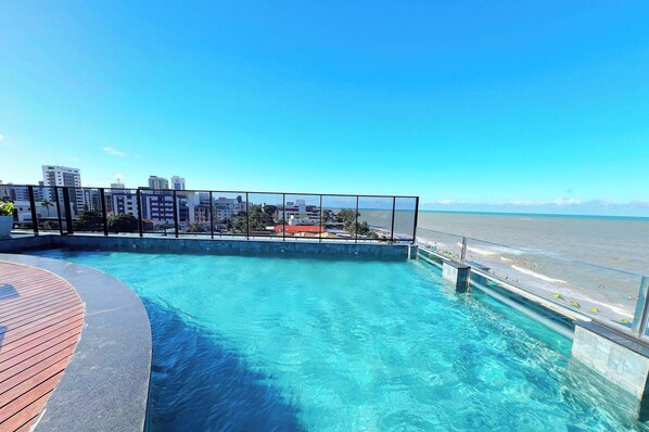 A swimming pool with a wooden deck, a glass railing, and a view of the beach and buildings. - Alta Marine - Miramar Hospedagens  (João Pessoa)
