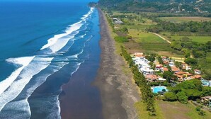 Una zona costera con una playa extensa, un pequeño poblado con casas y una piscina, y colinas al fondo.