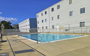 A swimming pool with a metal ladder, a fenced area, and a building with multiple windows and a door. - 2-bedroom Condo in OCMD Centrally Located, 2 Parking Spaces, Pool, and Grills (Ocean City)
