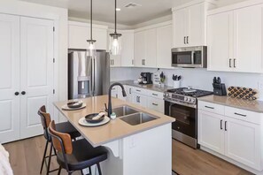 Una cocina moderna con armarios blancos, electrodomésticos de acero inoxidable, una isla central con taburetes altos e iluminación colgante.
