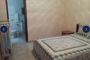 A bedroom with a bed, a nightstand, and a view of a bathroom with a toilet and a towel hanging on the wall.