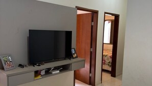 A modern living room with a flat-screen TV on a grey wall unit, a wooden door leading to another room, and a tiled floor. - Village house in Ponte Alta Babilônia - Serra da Canastra MG (Babilônia)