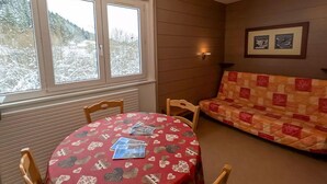 A cozy room with a round table covered in a red tablecloth with heart patterns, two wooden chairs, a red and beige sofa, and a window with a view of snow-covered trees.