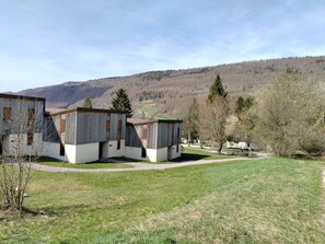 Um edifício rústico em estilo de cabana com telhado plano e uma pequena estrutura anexa, cercado por uma área gramada e árvores, com montanhas ao fundo.