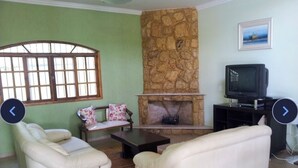 A living room with a stone fireplace, two white sofas, a television, and a window with a view. - Country house in Boituva, located in a gated community. Large swimming pool, Wi-Fi, small soccer field. (Boituva)