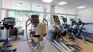 Un gimnasio con diversas máquinas de cardio, incluyendo cintas de correr y elípticas, y un amplio ventanal que ofrece vistas a palmeras y edificios.