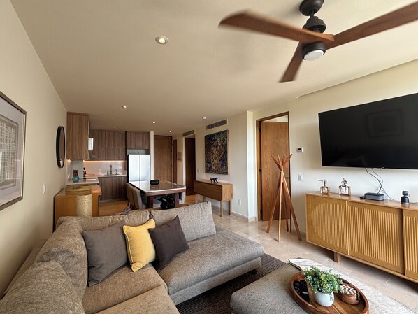 Una sala moderna con sofá, una mesa de centro y televisor. Se observa una zona de cocina con armarios y una mesa de comedor.