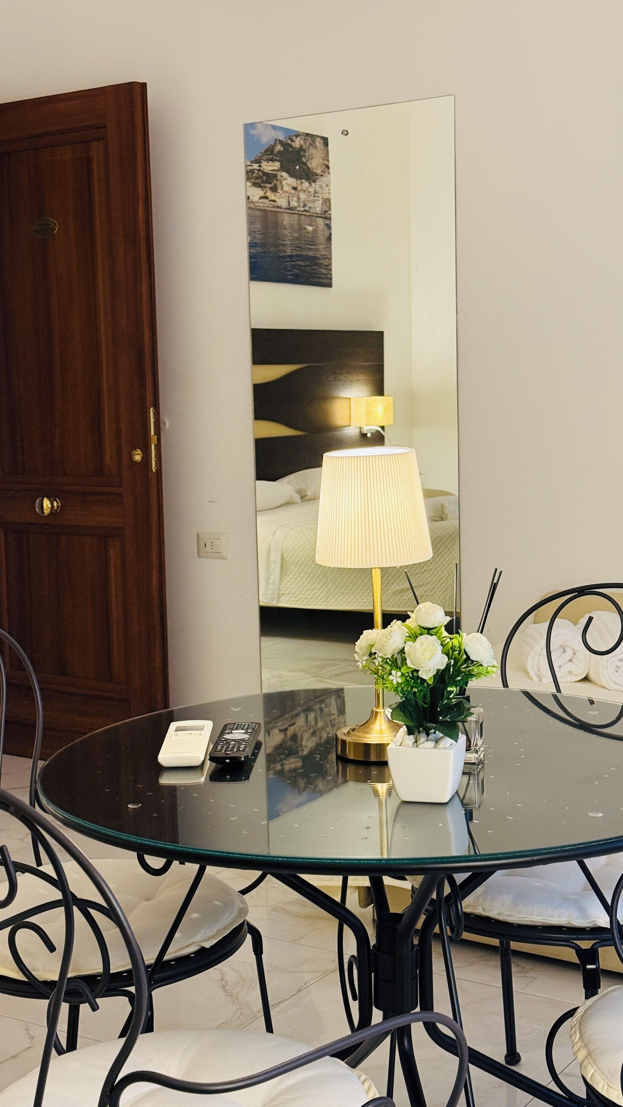 Una camera d'albergo con un tavolino rotondo in vetro, una porta in legno, un letto con lenzuola bianche, una lampada da comodino e uno specchio che riflette la stanza.