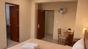 Et hotelværelse med en seng, et skrivebord, en stol, en ventilator og et lille bord med en plante.