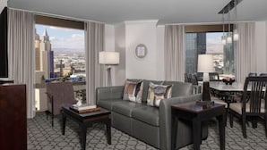 A modern hotel room with a grey sofa, a small table, and a dining area with a view of the cityscape.