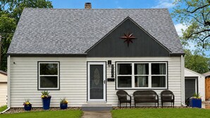 Ett envåningshus med ett mörkt tak, vit träpanel och en stjärnformad dekoration på framsidan. Det finns två fönster på framsidan, en dörr och en liten veranda med två stolar.