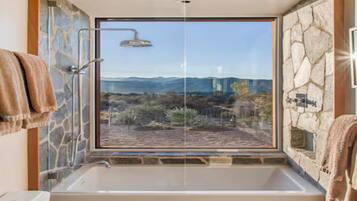 Un baño con una bañera grande, ducha y vistas a un paisaje montañoso a través de un ventanal amplio.
