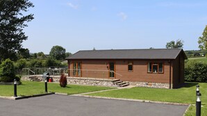 A small, single-story wooden building with a sloped roof, a stone foundation, and a paved entrance area. - Pine Lodge (Holywell)