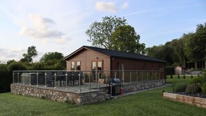A house with a stone foundation, a covered patio with glass railings, and a barbecue grill. - Pine Lodge (Holywell)
