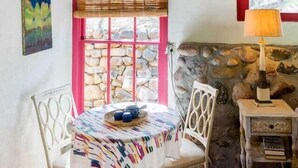 Un comedor con una mesa redonda, sillas, una pared de piedra, una ventana con persianas y una lámpara.