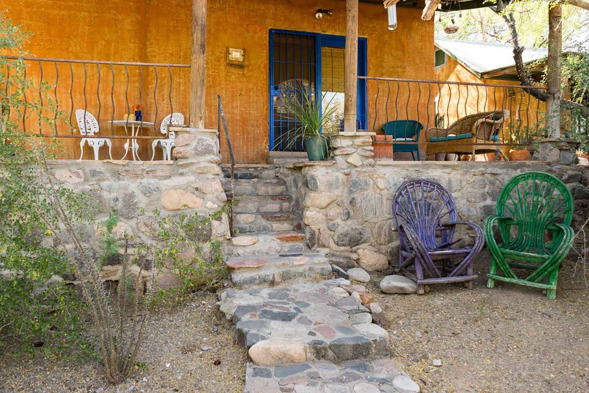 A rustic outdoor seating area with stone steps leading to a wooden door, surrounded by greenery and stone walls.