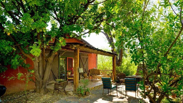 Un'area relax esterna rustica con pavimento in pietra e arredi in legno, immersa nel verde e adiacente a un edificio rosso.