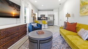 Un salon moderne avec un téléviseur à écran plat, une table basse ronde, un canapé jaune et une cuisine en arrière-plan.