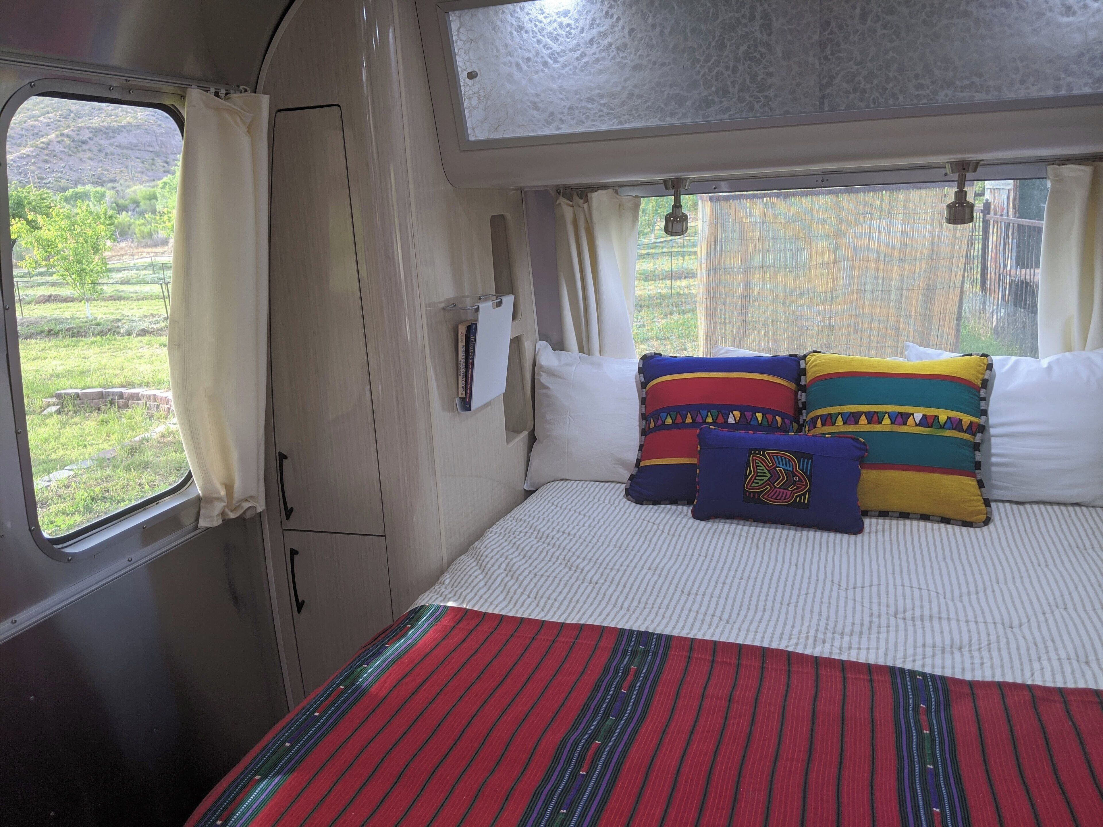 A bed with striped bedding and colorful pillows, a window with curtains, and a view of greenery outside.