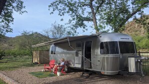 A silver Airstream RV parked in a grassy area with a red chair and a potted plant nearby.