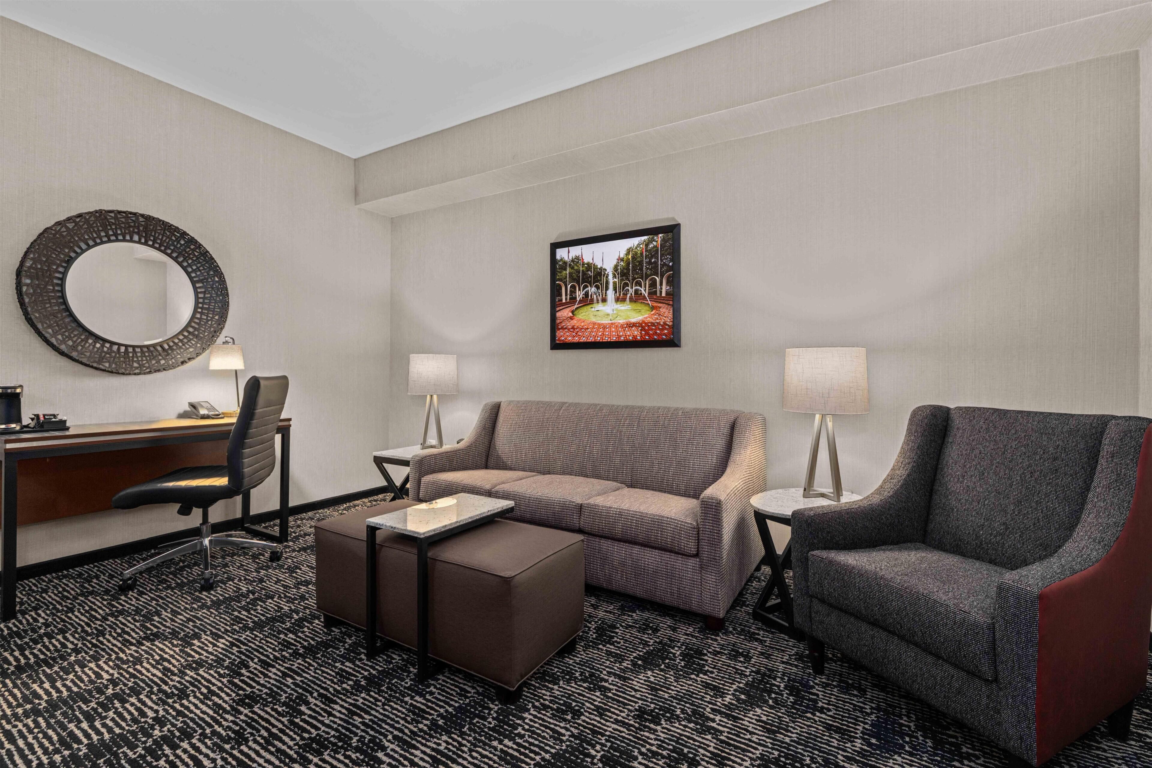 A hotel room with a desk, two chairs, a sofa, a small ottoman, a round mirror, two lamps, and a framed picture on the wall.