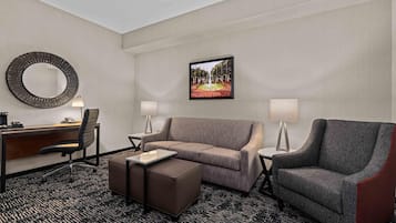 A hotel room with a desk, two chairs, a sofa, a small ottoman, a round mirror, two lamps, and a framed picture on the wall.