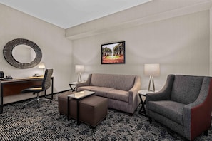 Habitación de hotel con escritorio, dos sillas, sofá, pequeño reposapiés, espejo redondo, dos lámparas y un cuadro en la pared.