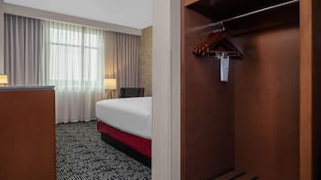Quarto de hotel com cama, escrivaninha, cadeira, guarda-roupa e janela com cortinas.