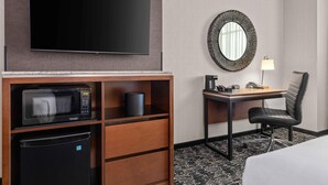 Habitación de hotel con televisión de pantalla plana, microondas, refrigerador, escritorio con silla, lámpara y un espejo decorativo.