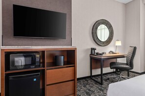 Habitación de hotel con televisión de pantalla plana, microondas, refrigerador, escritorio con silla, lámpara y un espejo decorativo.