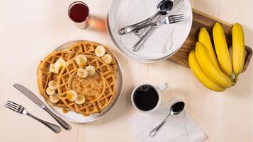 A breakfast setting with waffles, bananas, syrup, coffee, and cutlery on a wooden table.