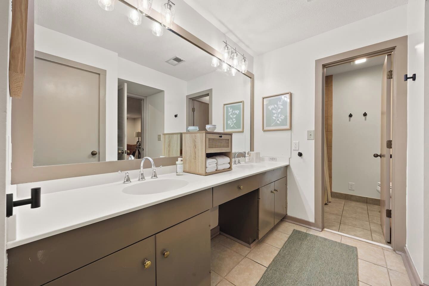 Une salle de bain moderne avec un grand meuble-lavabo, un double vasque et un grand miroir. Des tableaux encadrés ornent le mur, et un meuble en bois se trouve sous le miroir.