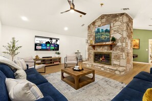 A modern living room with a stone fireplace, a mounted TV, and a ceiling fan. - Modern Family Retreat - Private Pool/Mini Golf/Fire Pit/Pool Table - Sleeps 14 (San Antonio)