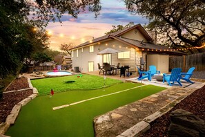 A backyard with a small artificial turf golf course, a wooden deck, and a house with string lights. - Modern Family Retreat - Private Pool/Mini Golf/Fire Pit/Pool Table - Sleeps 14 (San Antonio)