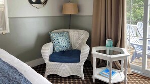 A room with a wicker chair, a small table with a glass top, a lamp, and a view of outdoor chairs through a window.