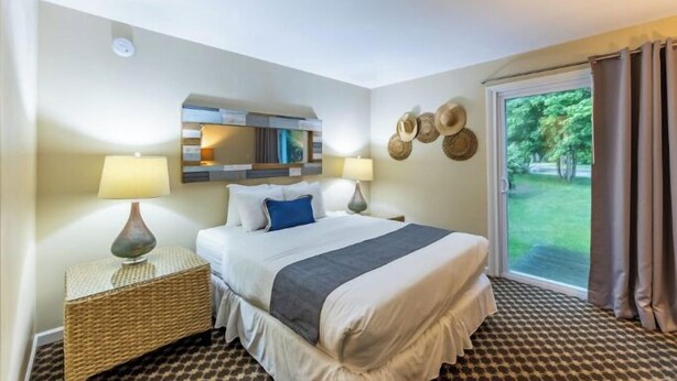 Habitación de hotel con cama, dos mesitas de noche con lámparas, una mesita auxiliar de mimbre, un espejo y vista al exterior a través de una puerta corrediza de vidrio.