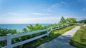 A coastal walkway with a white fence, grassy areas, and a clear view of the ocean.