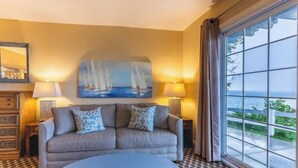 A room with a sofa, two lamps, a painting of sailboats, and a large window with a view of the sea.