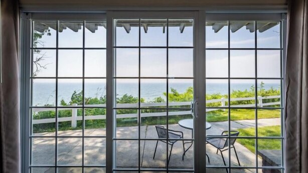 A sliding glass door with a view of a patio area with a round table and two chairs, a white fence, and a grassy landscape.