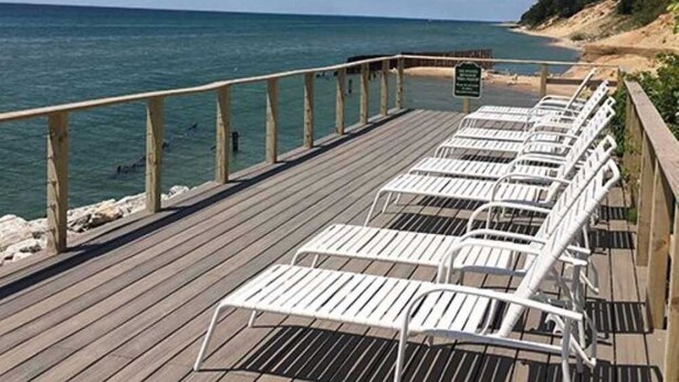 Eine Holzterrasse mit weißen Liegestühlen, die zum Meer ausgerichtet sind, einem Holzgeländer und einem Hinweisschild.