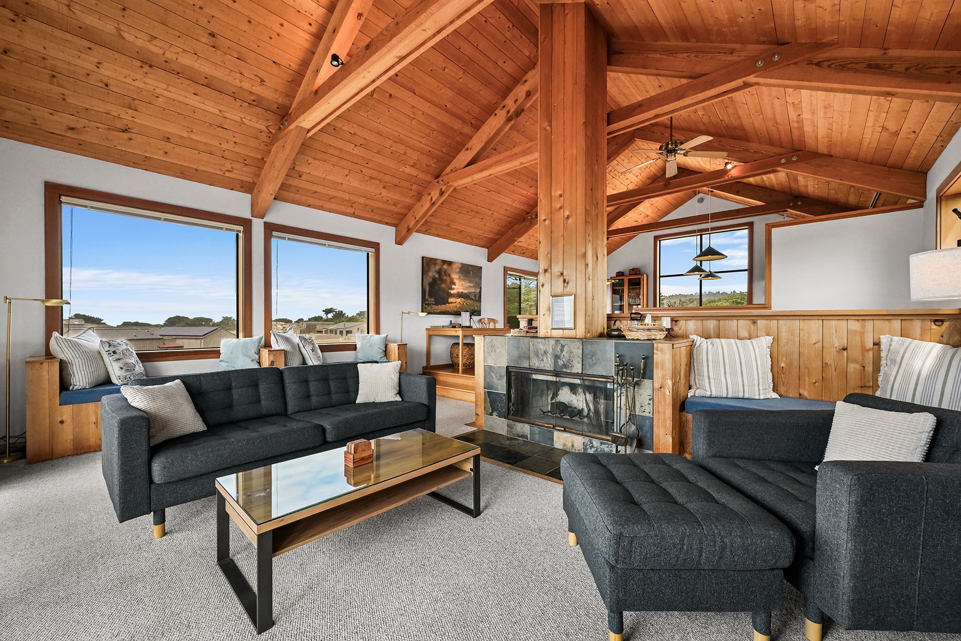A spacious living room with a fireplace, large windows, and a high wooden ceiling.