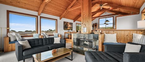 A spacious living room with a fireplace, large windows, and a high wooden ceiling.