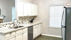 Cocina con armarios blancos, encimera de granito, refrigerador de acero inoxidable y ventana con persianas.