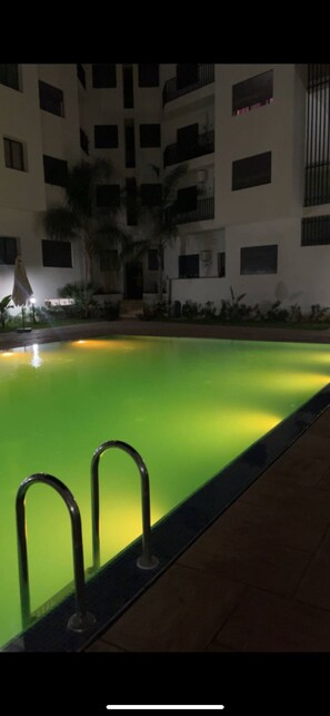 A nighttime view of a swimming pool with a green hue, surrounded by a multi-story building with balconies. - Apartment with a pool, a large terrace, and a balcony overlooking the pool. (Bouznika)