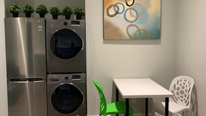 Una cocina compacta con refrigerador de acero inoxidable, lavadora y secadora integradas, una mesa blanca y una silla verde.