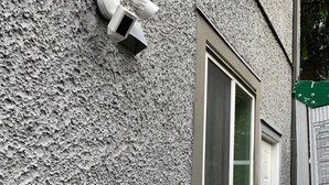 Fachada de edificio con pared de estuco texturizado, una ventana blanca con marco blanco y una cámara de seguridad montada en la pared.