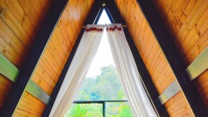 A room with a bed, red bedding, white curtains, and a window with a view of greenery. - El Encanto de Ráquira Lodge (raquira)
