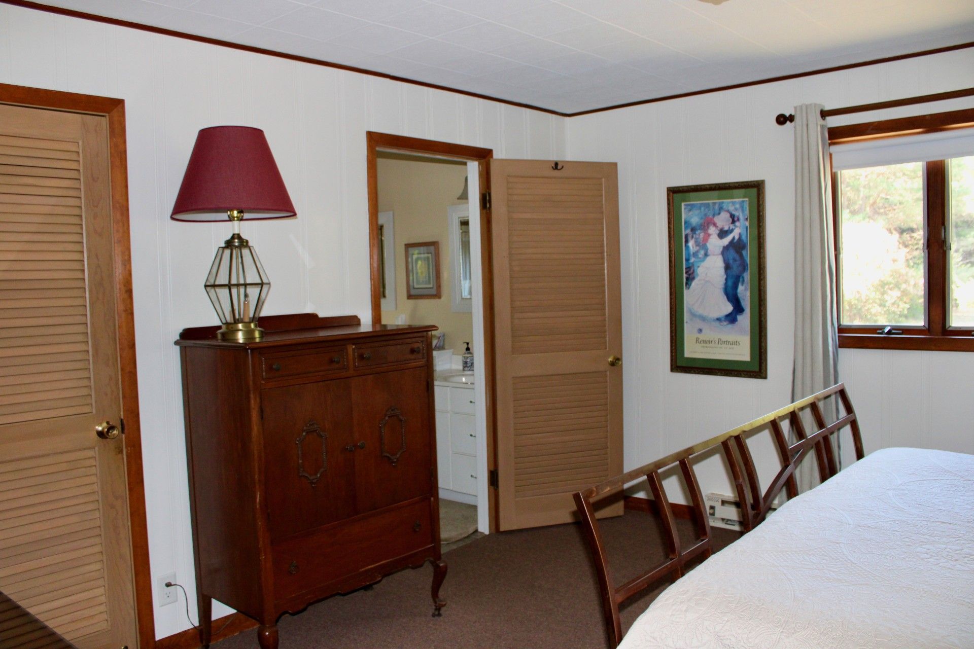 A bedroom with a wooden dresser, a lamp, a framed picture, and a window.